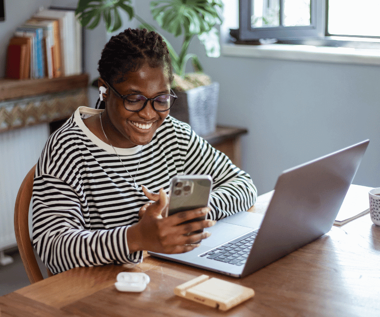 A social media freelancer sits at a desk working on their mobile phone with a laptop and earphones nearby