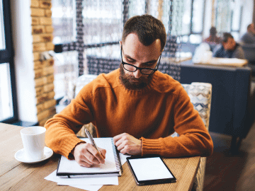 A male freelance writer sits writing on a notepad in a coffee shop