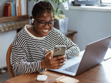 A social media freelancer sits at a desk working on their mobile phone with a laptop and earphones nearby