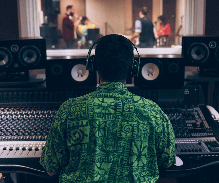 Male sound engineer sits at a recording desk with headphones on. Musicians are visible in the recording booth