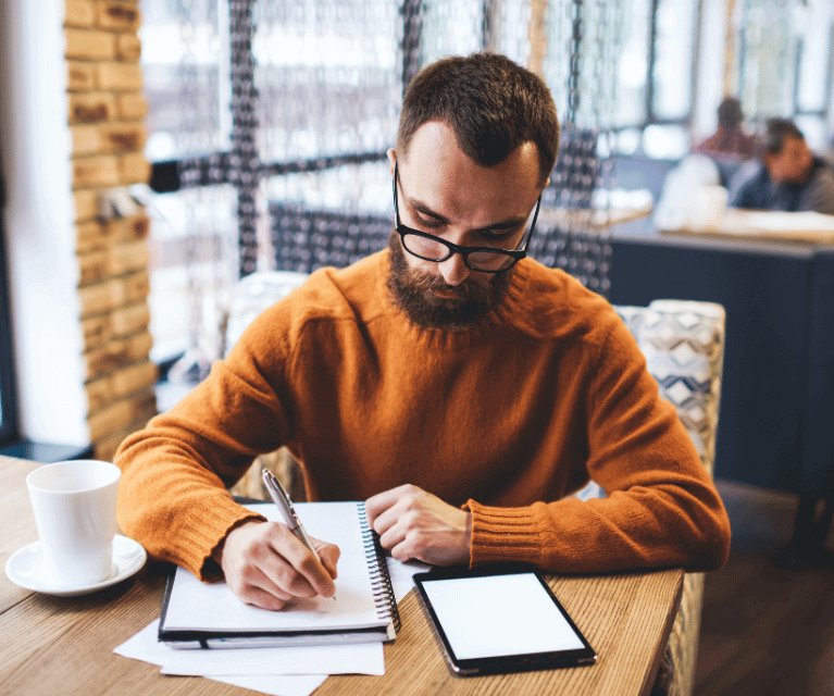 A male freelance writer sits writing on a notepad in a coffee shop