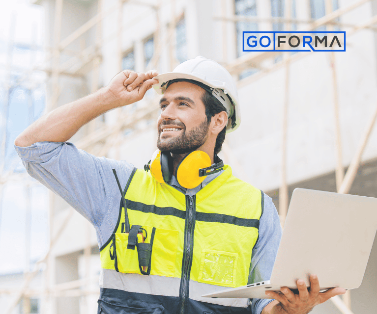 Foreman on a construction site holding a laptop and wearing a hard hat, high visibility vest. A walkie talkie is also visible.
