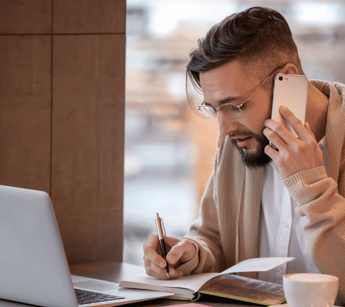 Photo of male freelancer using a mobile phone, sat in front of a laptop while making notes in a notebook